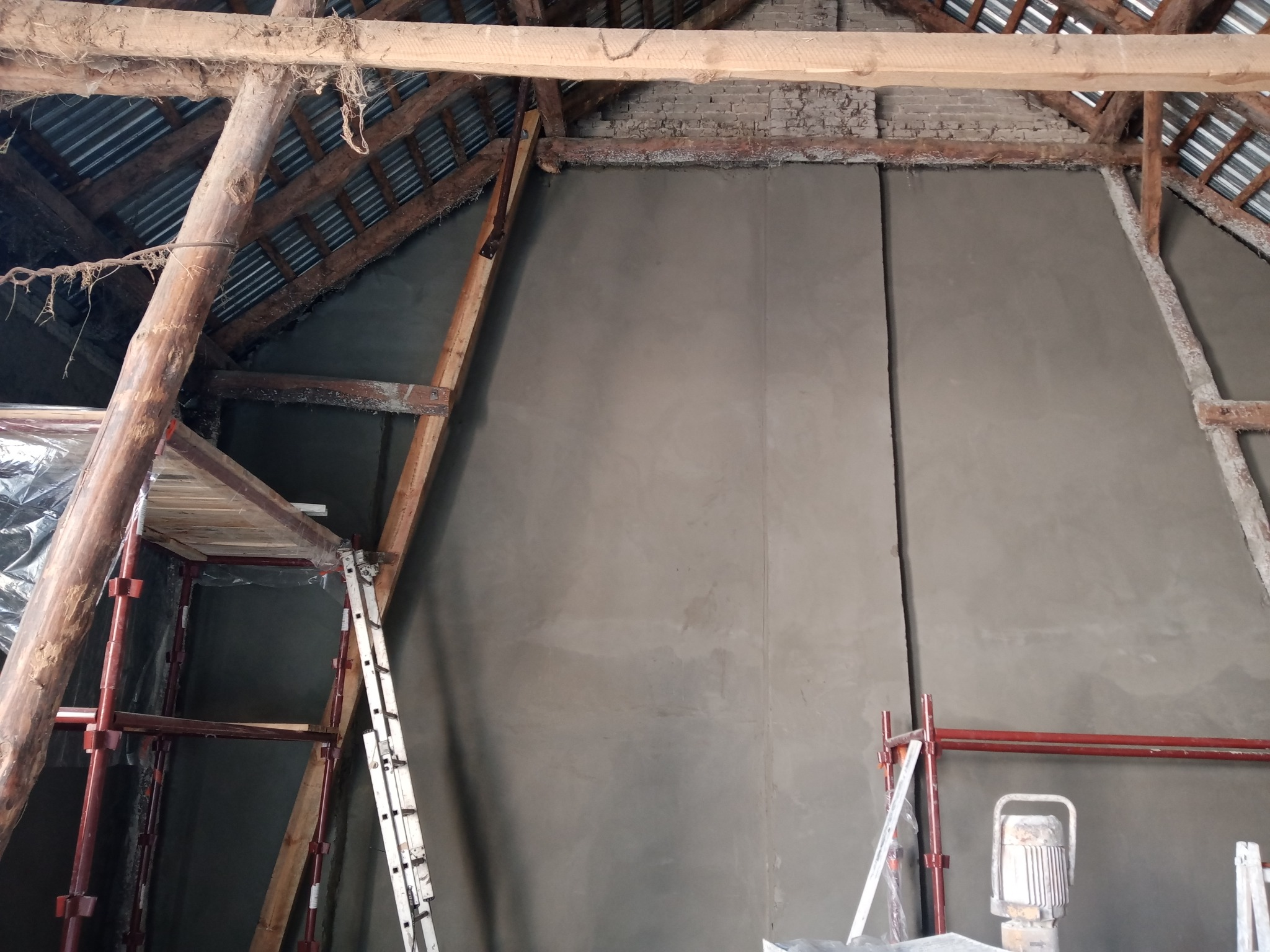 Świeżo otynkowana ściana poddasza z widoczną konstrukcją drewnianą więźby dachowej, rusztowaniem i drabiną w trakcie prac wykończeniowych.