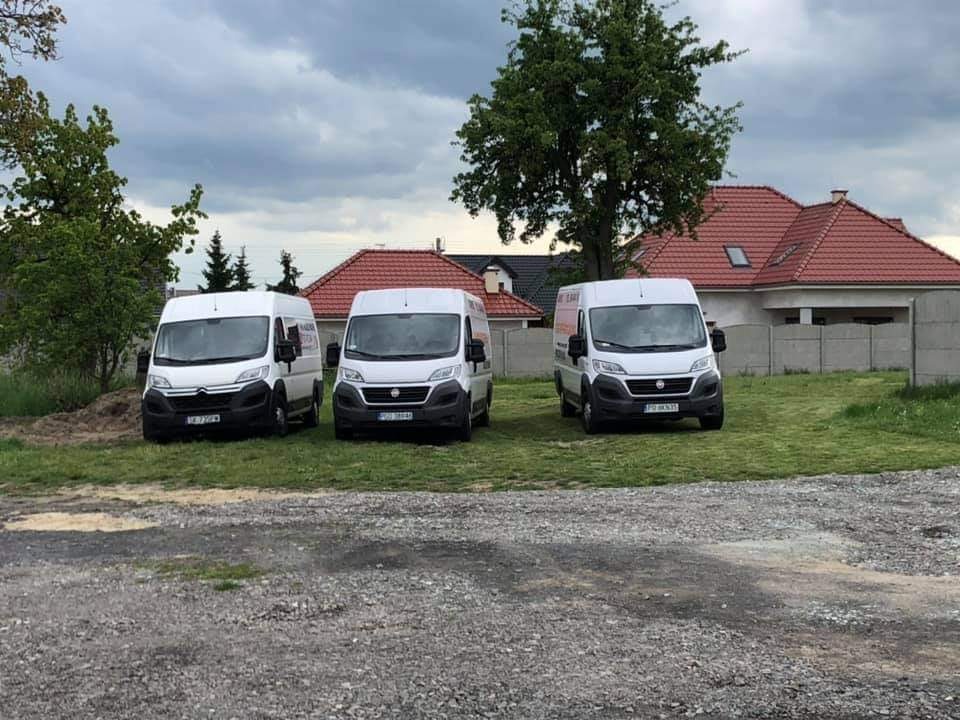 Trzy białe busy dostawcze zaparkowane na trawiastym terenie przed domem z czerwoną dachówką w pochmurny dzień.