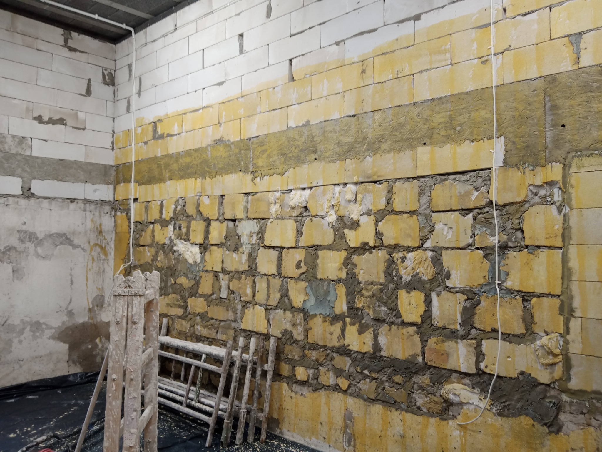 Wnętrze w trakcie prac murarskich: ściana z częściowo otynkowanych bloczków, widoczne warstwy izolacji i fragmenty starej farby, drewniana drabina oparta o ścianę.