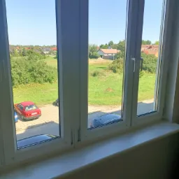 Widok z okna PCV na podwórko z czerwonym i niebieskim samochodem w słoneczny dzień.