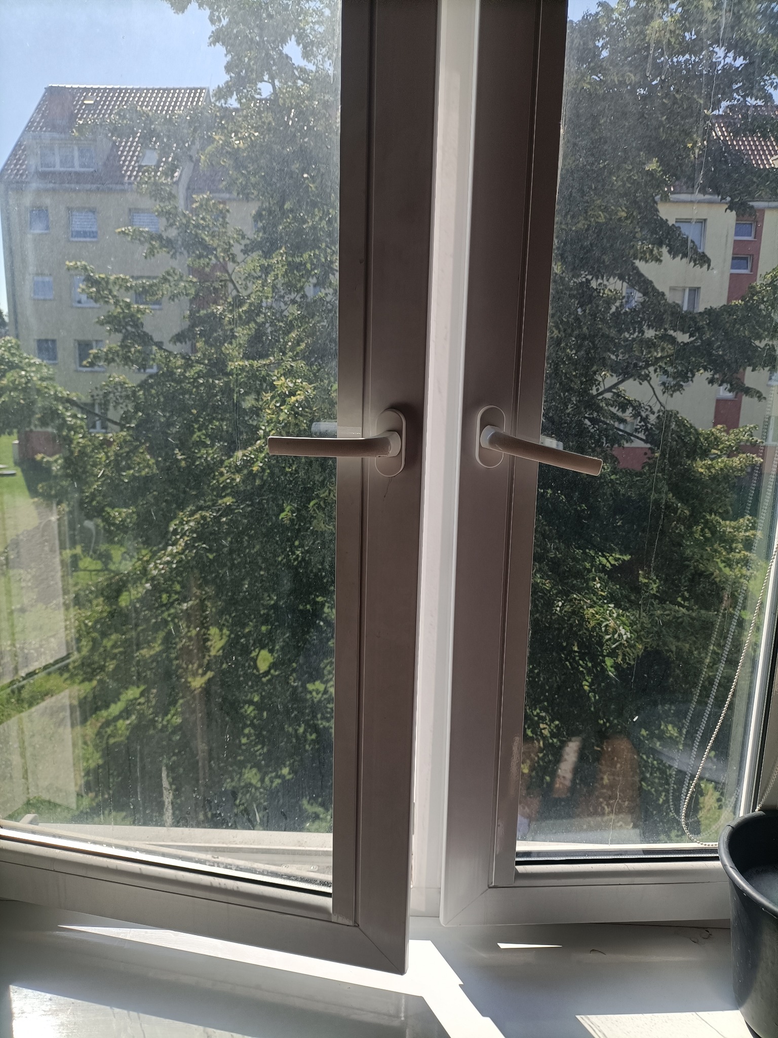 Okno PCV z widocznymi smugami i zaciekami na szybie, przez które widać budynek i zieleń drzew, uchwycone z bliska przed czyszczeniem.