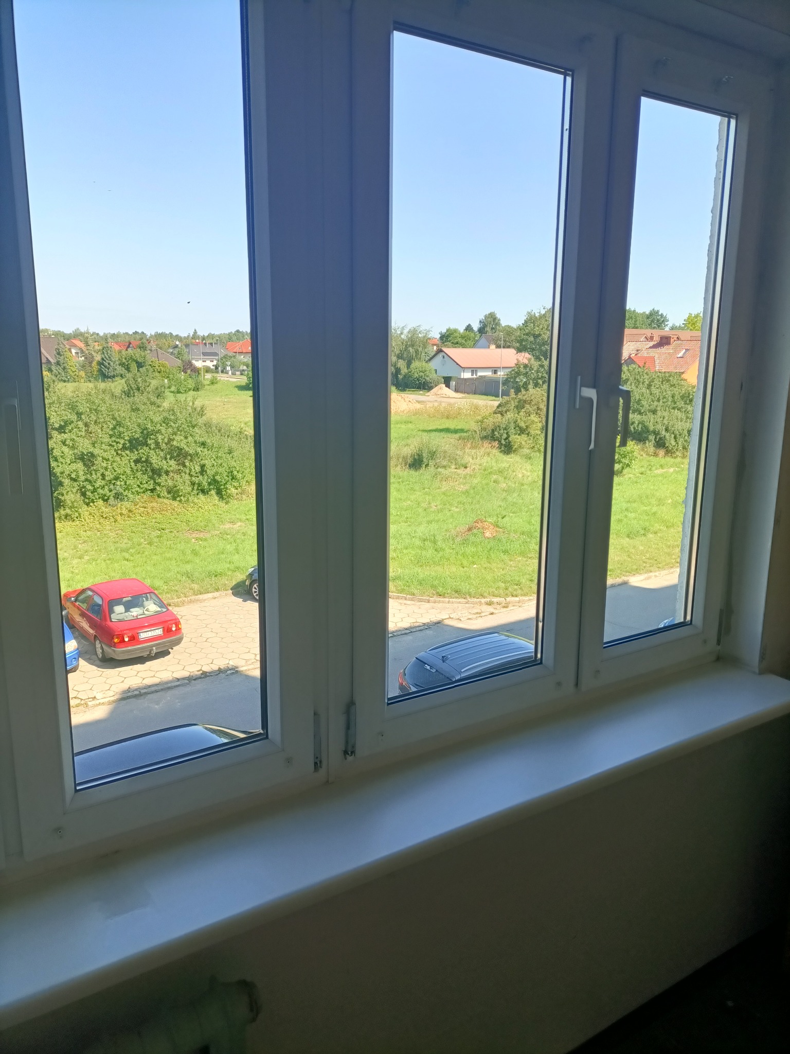 Widok z okna PCV na podwórko z czerwonym i niebieskim samochodem w słoneczny dzień.