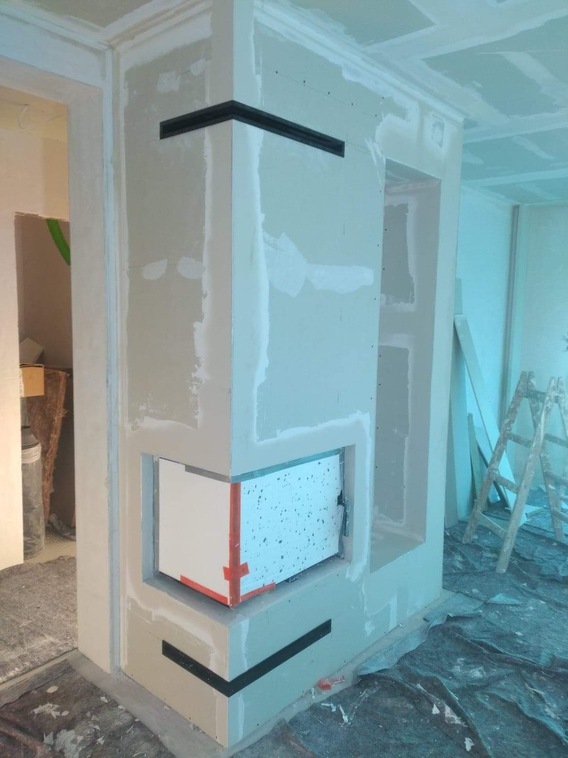 Wnętrze w trakcie prac wykończeniowych, widoczna zabudowa kominka z płyt gipsowo-kartonowych, zabezpieczona folią i taśmą, z elementami dekoracyjnymi w kolorze czarnym. Na podłodze rozłożona folia...