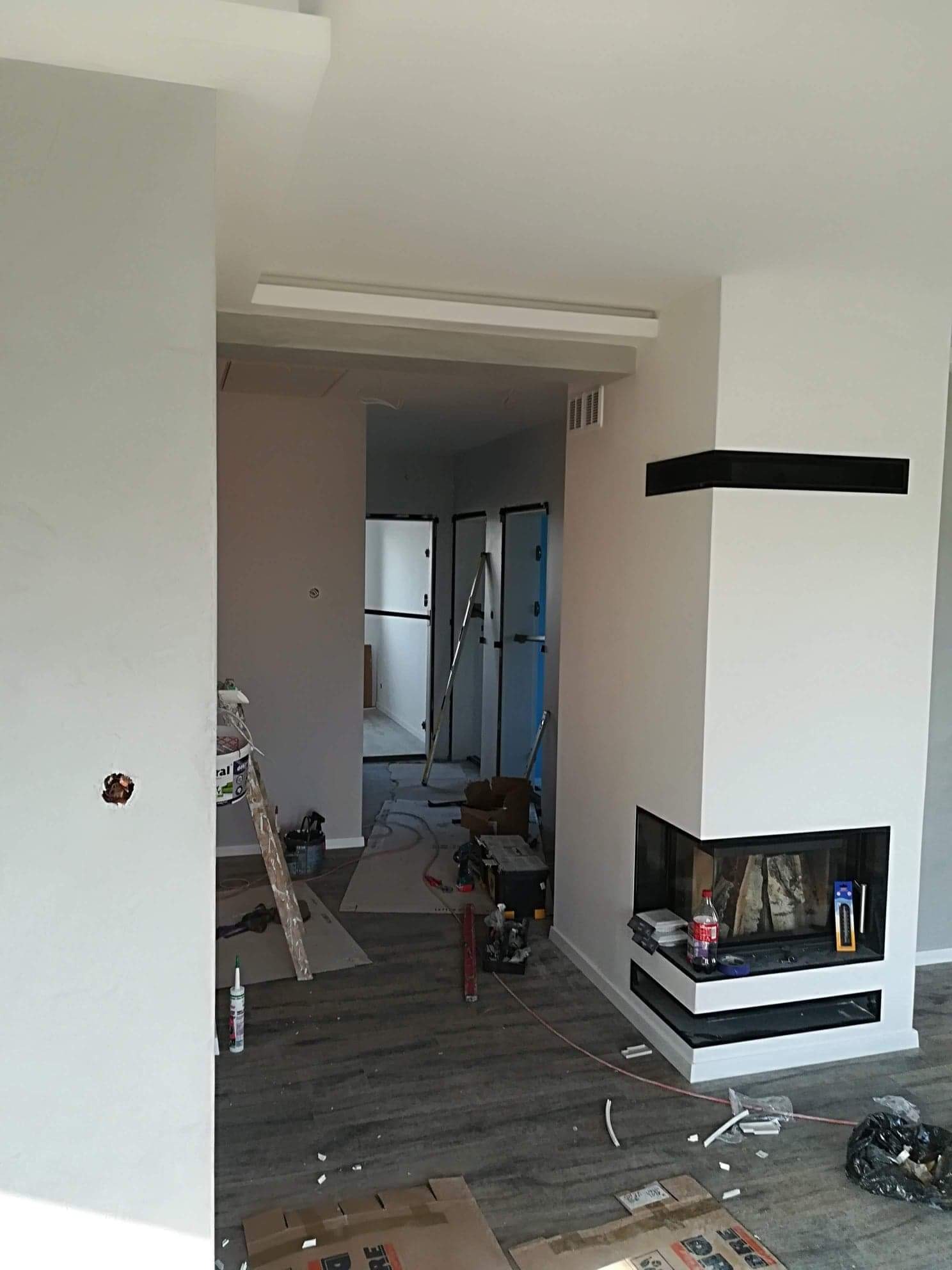 Wnętrze w trakcie prac wykończeniowych: szpachlowana ściana z widocznym otworem na gniazdko, drabina malarska, nowoczesny kominek narożny z czarnymi elementami, podłoga z paneli imitujących drewno,...