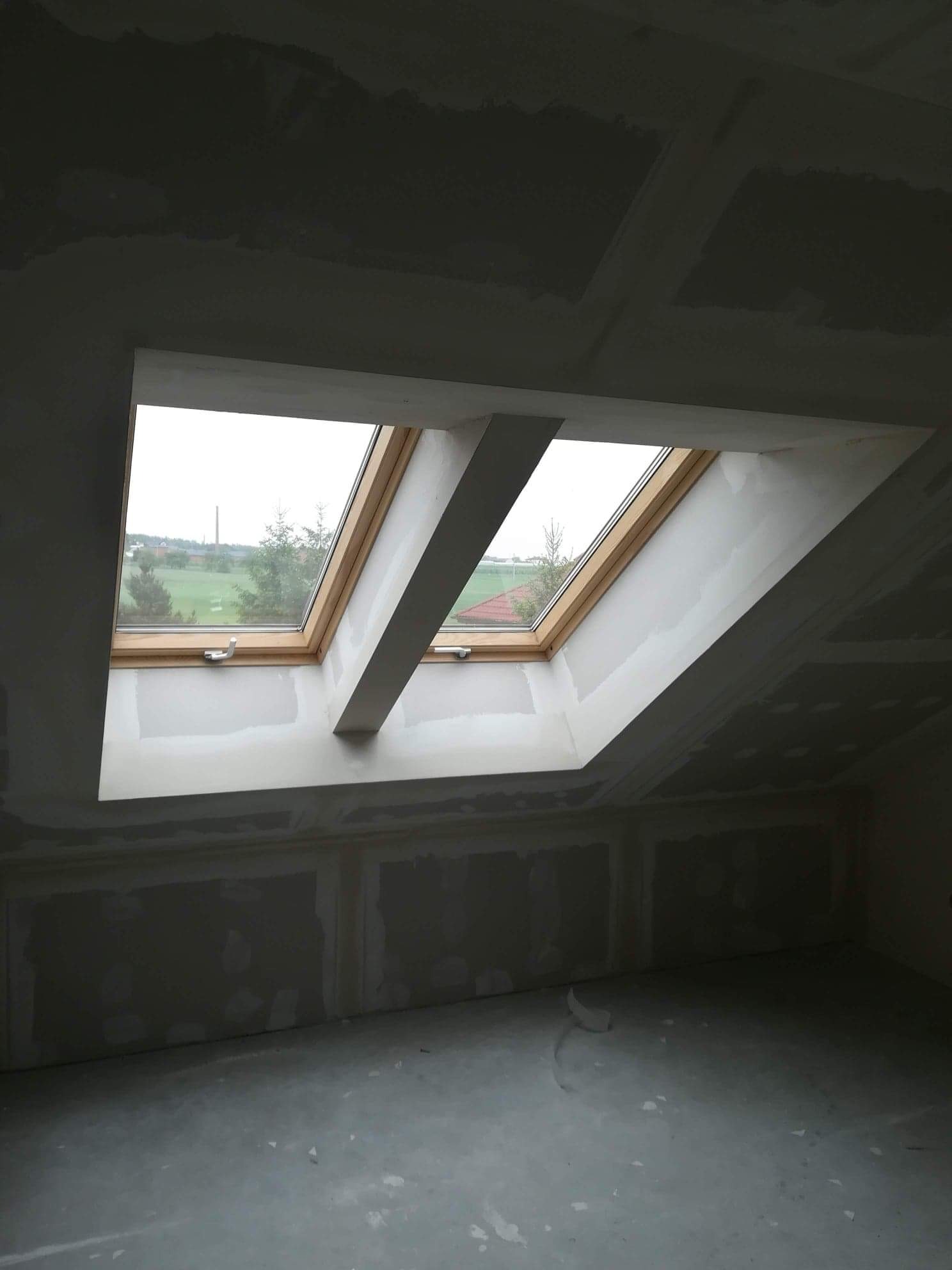 Poddasze w trakcie ocieplania z dwoma oknami dachowymi z drewnianymi ramami, widok na zielone pola i komin w tle, ściany i sufit pokryte płytami gipsowo-kartonowymi przed szpachlowaniem.