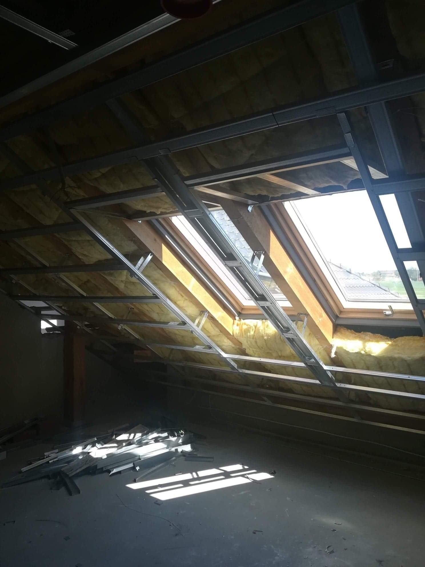 Poddasze w trakcie ocieplania, widoczne okna dachowe, wełna mineralna między krokwiami i metalowa konstrukcja nośna, na podłodze resztki profili.