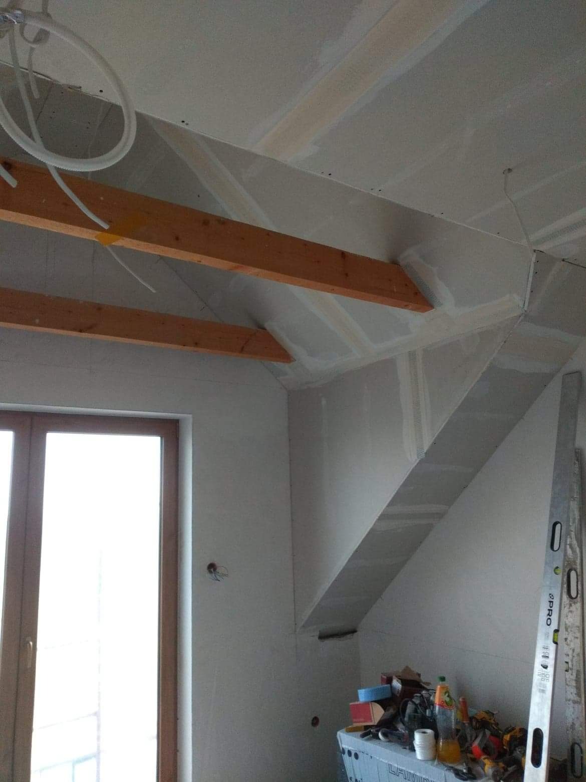 Poddasze w trakcie adaptacji, widoczne drewniane belki, instalacja elektryczna, płyty gipsowo-kartonowe i narzędzia na stole warsztatowym.