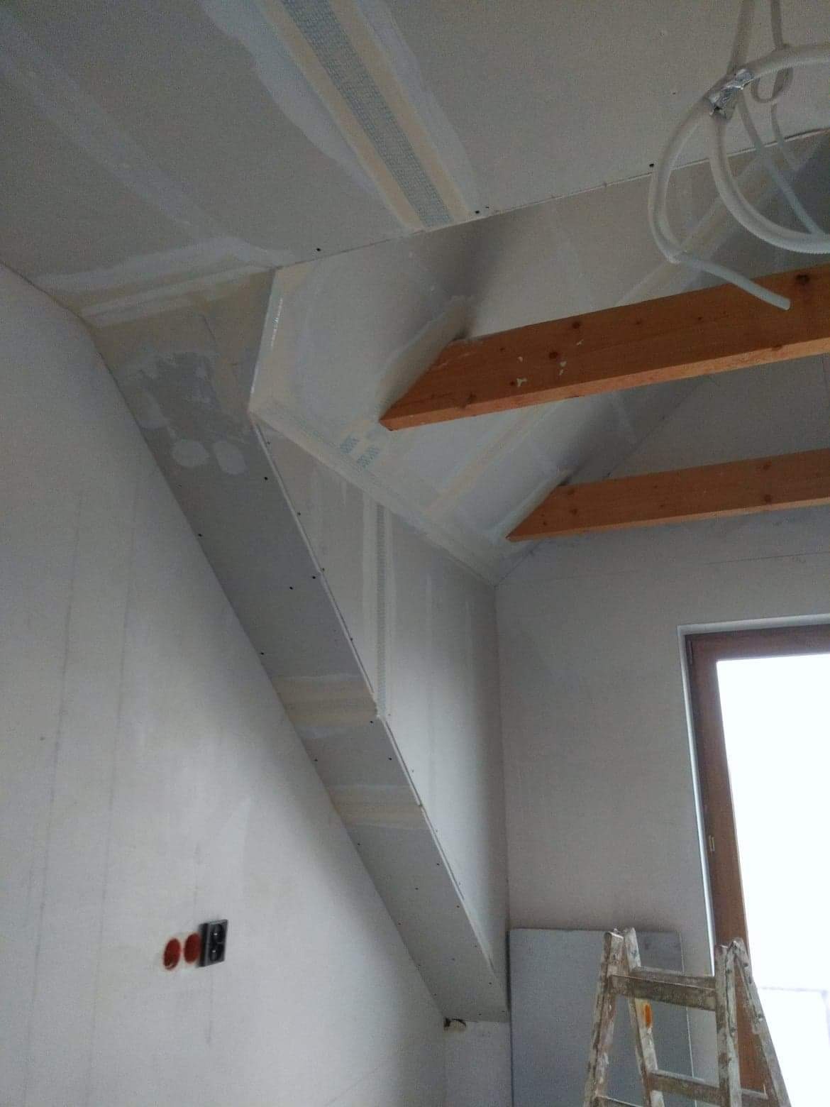 Poddasze w trakcie zabudowy płytami gipsowo-kartonowymi, widoczne drewniane elementy konstrukcyjne, instalacja elektryczna i drabina oparta o ścianę przy oknie.