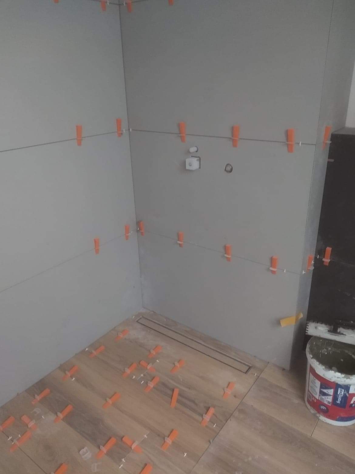 Montaż płytek imitujących drewno w kabinie prysznicowej z odpływem liniowym, widoczne pomarańczowe krzyżyki dystansowe i wiadro z klejem w rogu.
