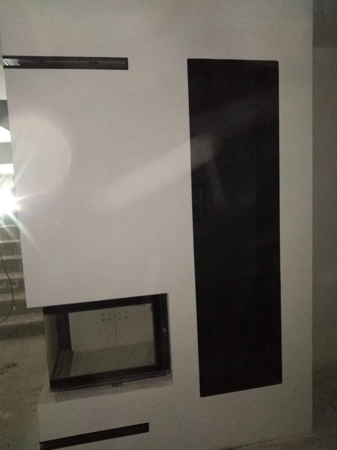 Nowoczesny kominek z białym obramowaniem i czarnymi akcentami, w trakcie montażu, widoczne niedoskonałości wykończenia ściany i podłogi.