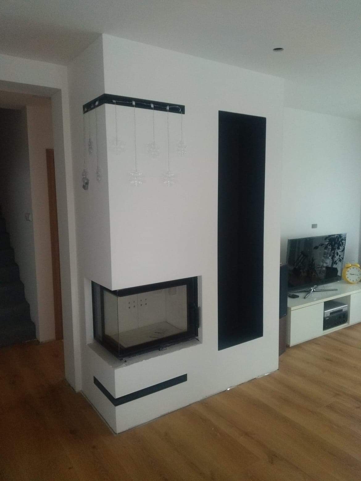 Nowoczesny kominek narożny z czarnymi akcentami w jasnym wnętrzu, ozdobiony wiszącymi dekoracjami w kształcie płatków śniegu, widok na telewizor i drewnianą podłogę.