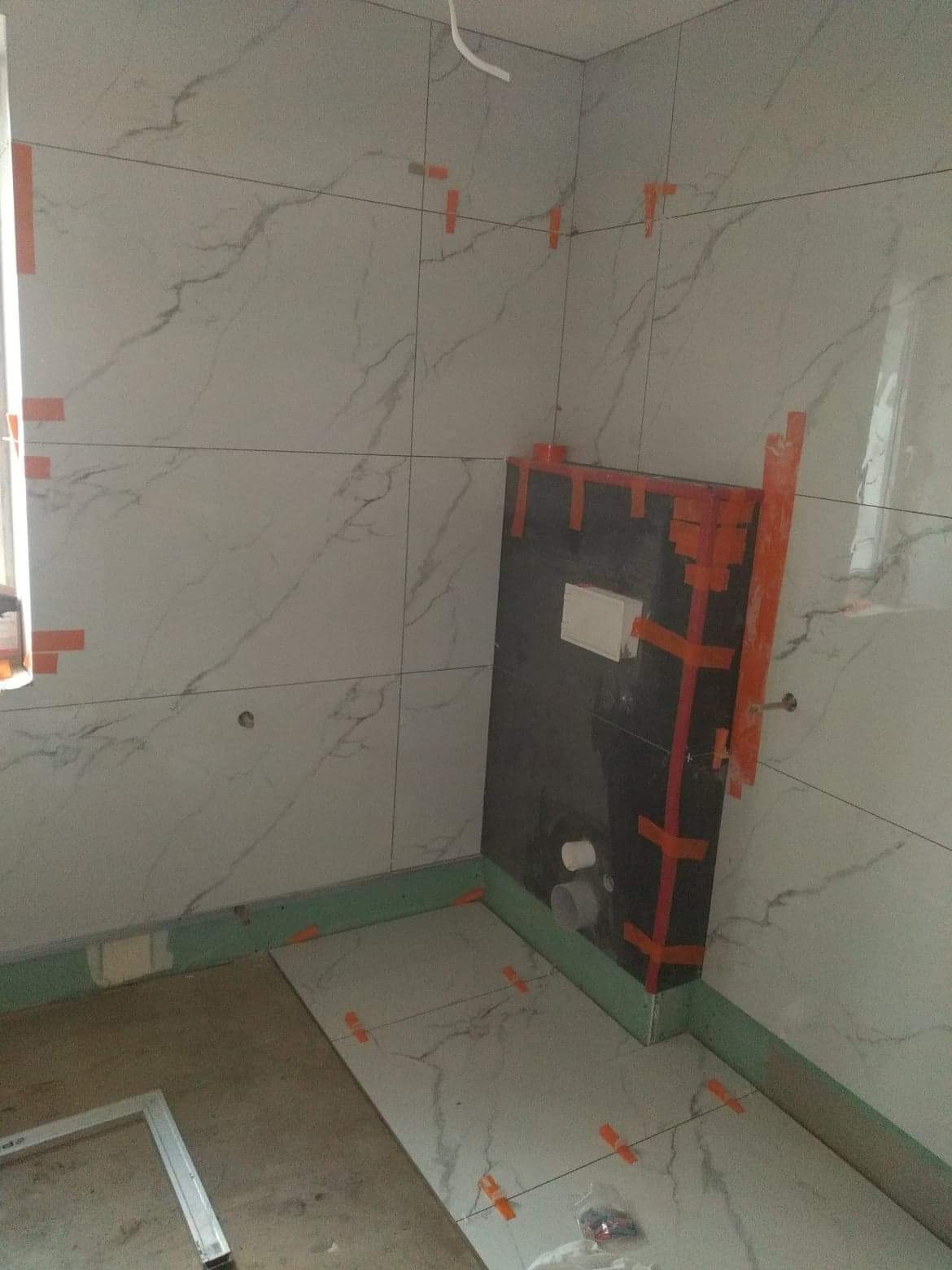 Łazienka w trakcie układania płytek imitujących marmur na ścianach i podłodze, z widocznym stelażem podtynkowym obłożonym czarną płytą i zabezpieczonym pomarańczową taśmą, oraz poziomą zieloną...