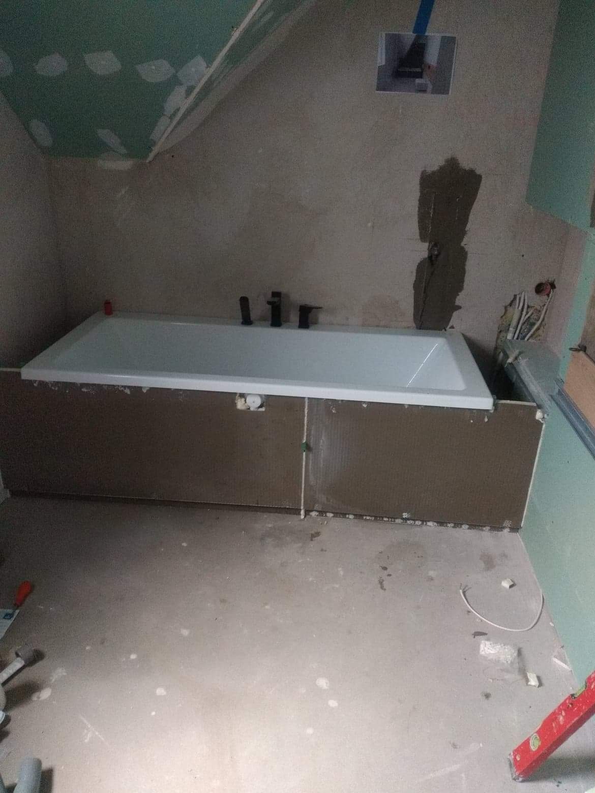 Wanna obudowana brązowymi płytami w surowej łazience w trakcie prac wykończeniowych, widoczne narzędzia na podłodze, ściany częściowo pokryte płytami gipsowo-kartonowymi, instalacja elektryczna...