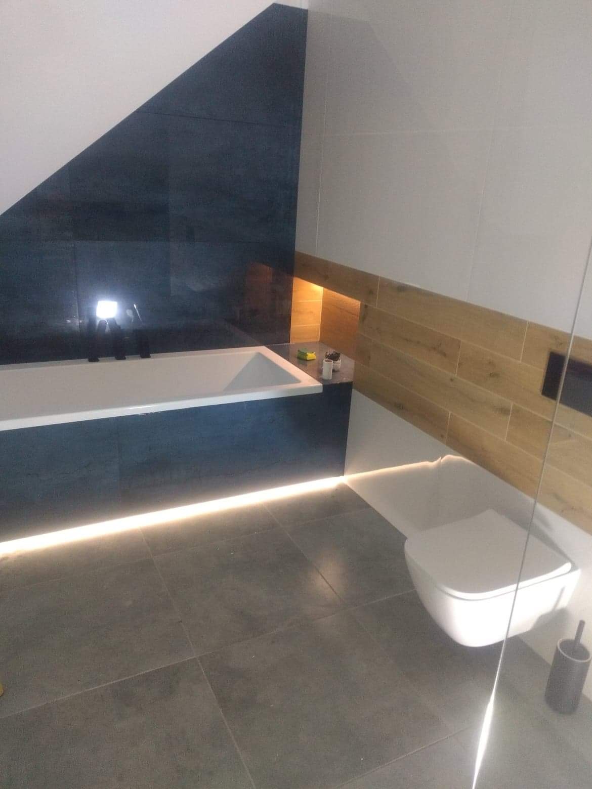 Nowoczesna łazienka w szaro-białej kolorystyce z podświetlaną wanną, drewnopodobnymi panelami i podwieszaną toaletą.