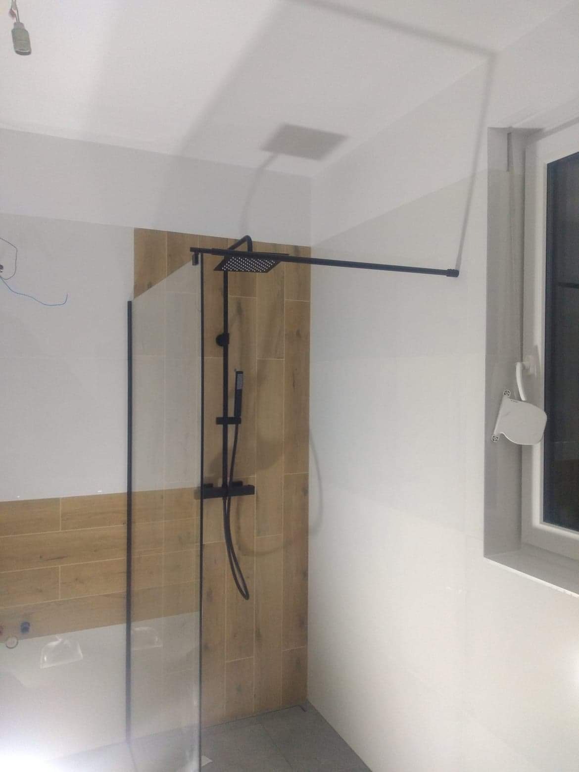 Kabina prysznicowa typu walk-in z czarną armaturą na tle ściany wyłożonej płytkami imitującymi drewno, widok na fragment łazienki z oknem.