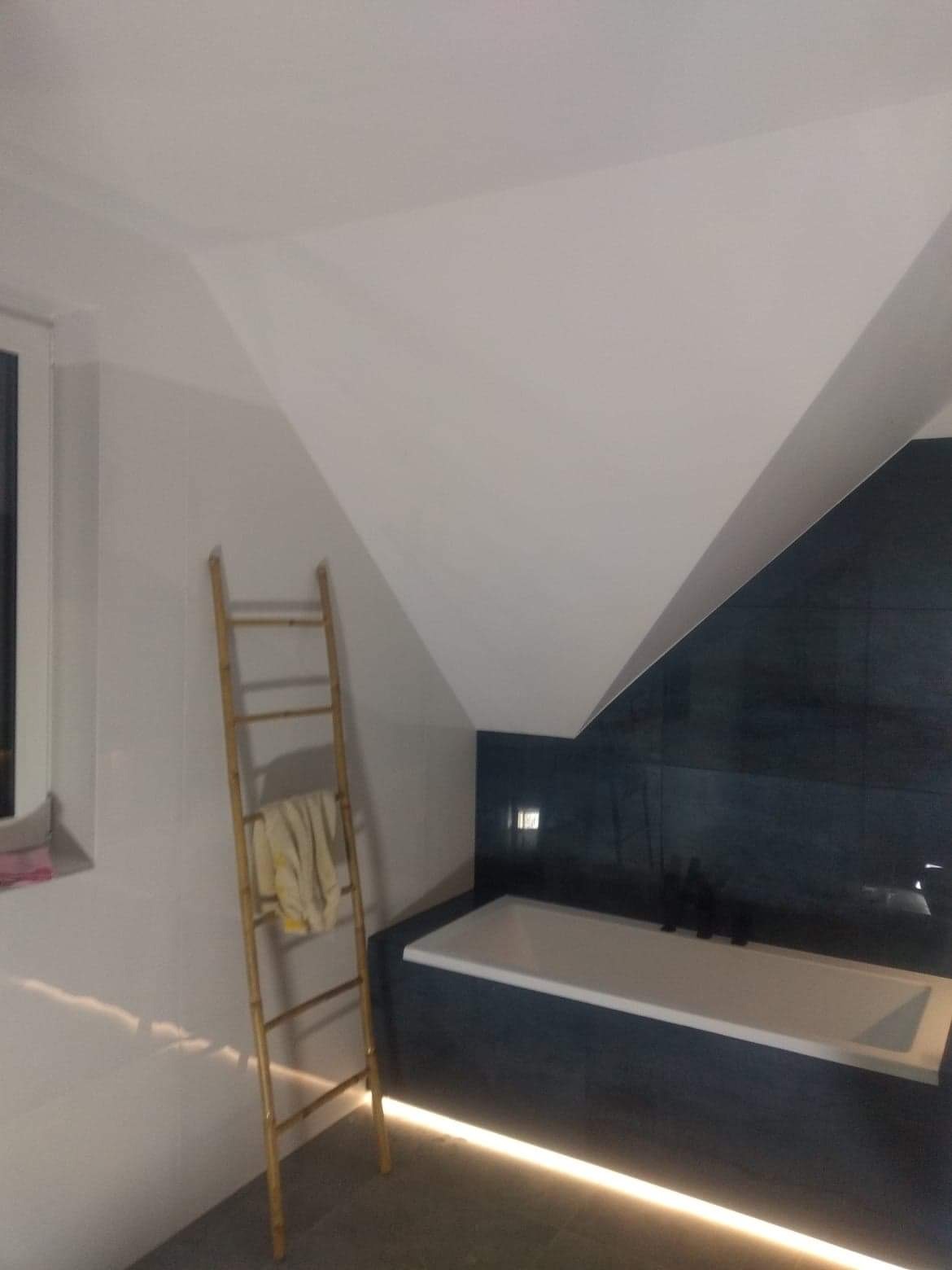 Nowoczesna łazienka na poddaszu z białą wanną w obudowie z ciemnych płytek, oświetleniem LED pod wanną i bambusową drabinką na ręczniki.