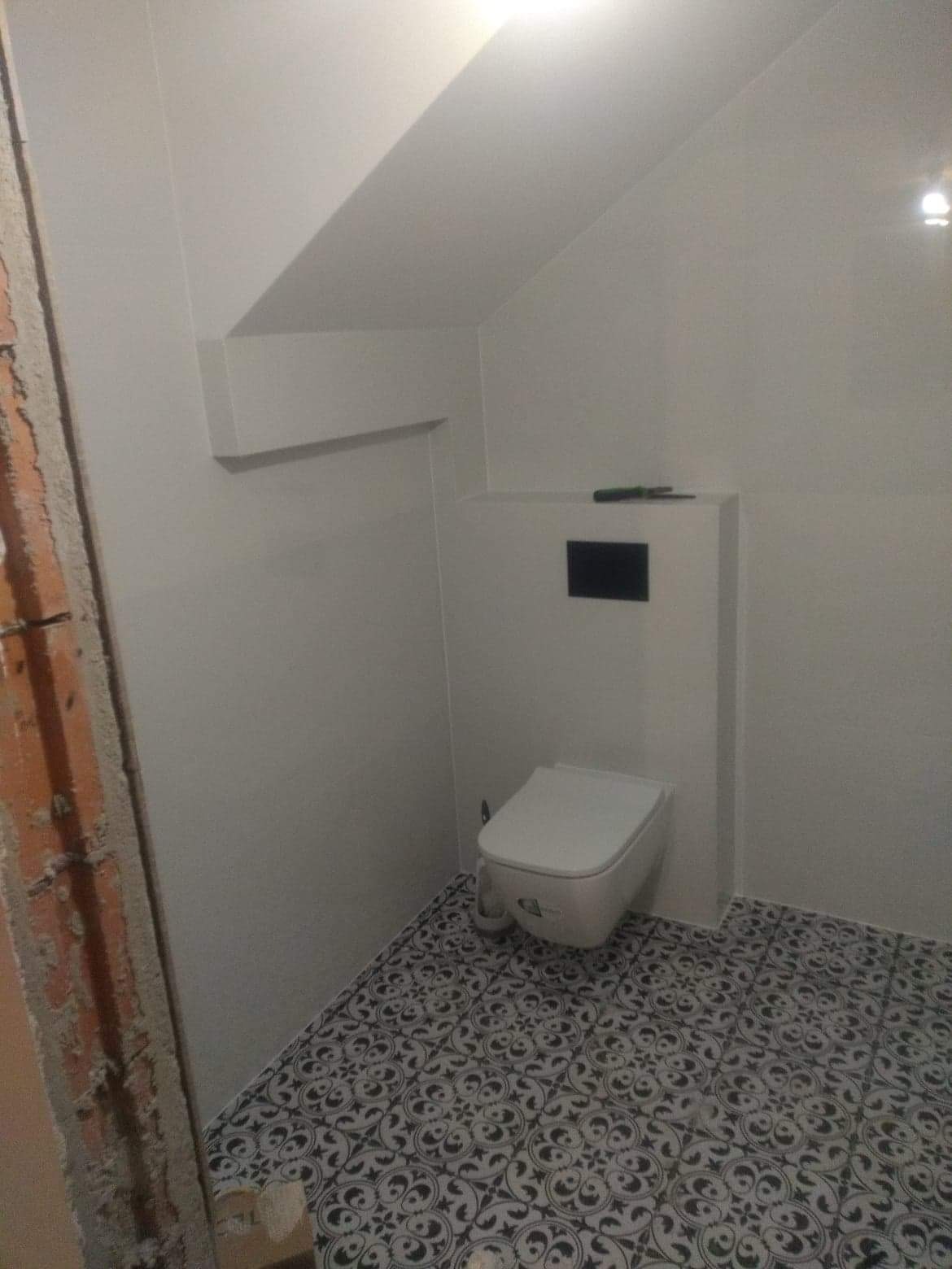 Nowoczesna, biała toaleta wisząca z czarnym przyciskiem spłukującym na tle białych płytek i wzorzystej, czarno-białej podłogi w łazience w trakcie remontu, widoczna surowa ściana z cegły.