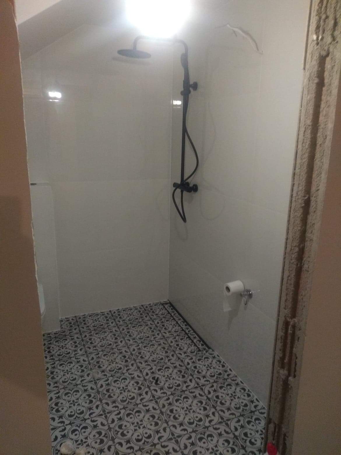 Nowoczesna kabina prysznicowa z czarną armaturą i wzorzystą podłogą, widoczny fragment nie wykończonego otworu drzwiowego.