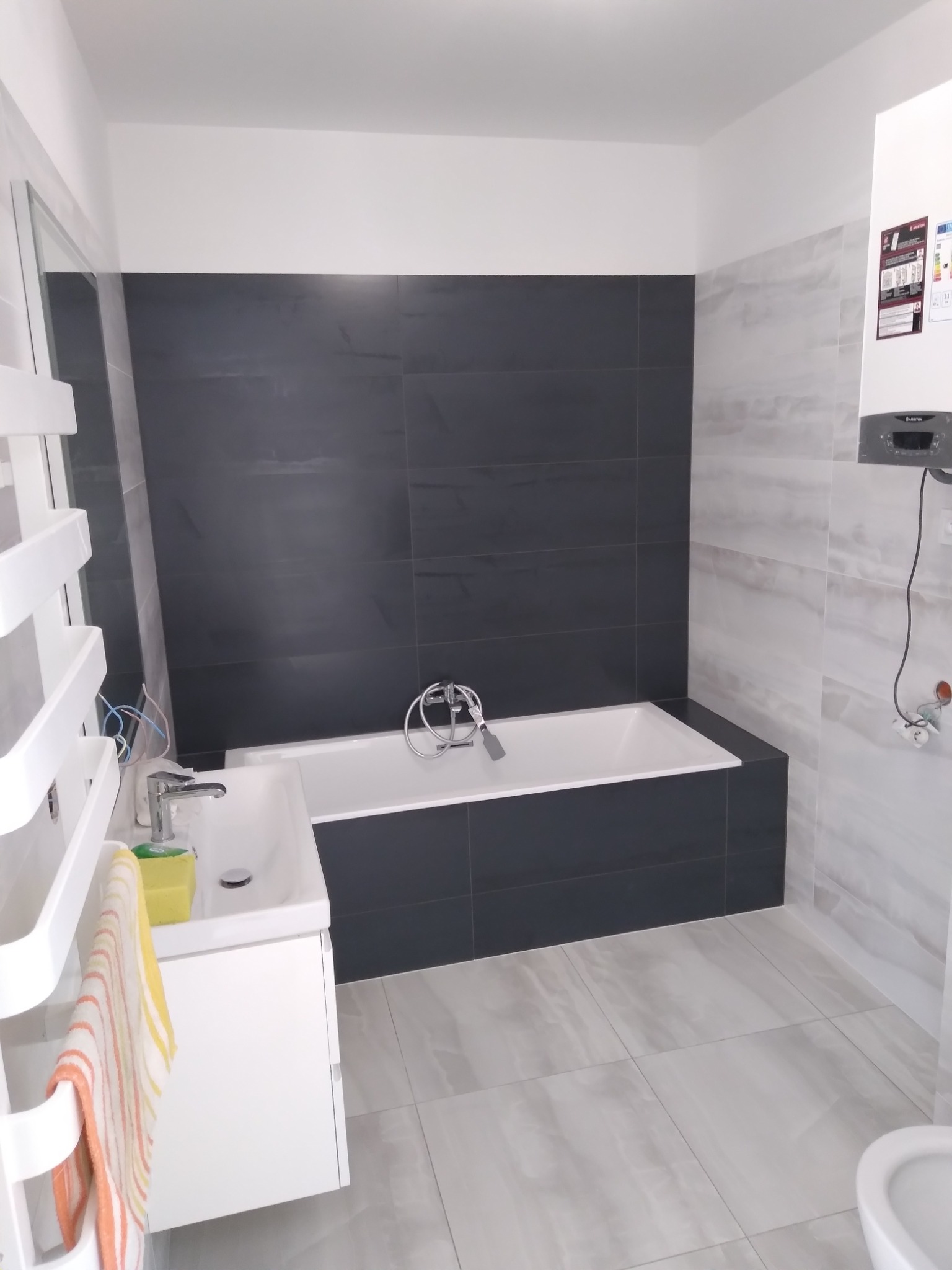 Nowoczesna łazienka z wanną obudowaną ciemnymi płytkami, jasnymi płytkami imitującymi marmur na ścianach i podłodze, oraz białymi meblami łazienkowymi i grzejnikiem drabinkowym z podłączonymi kablami.
