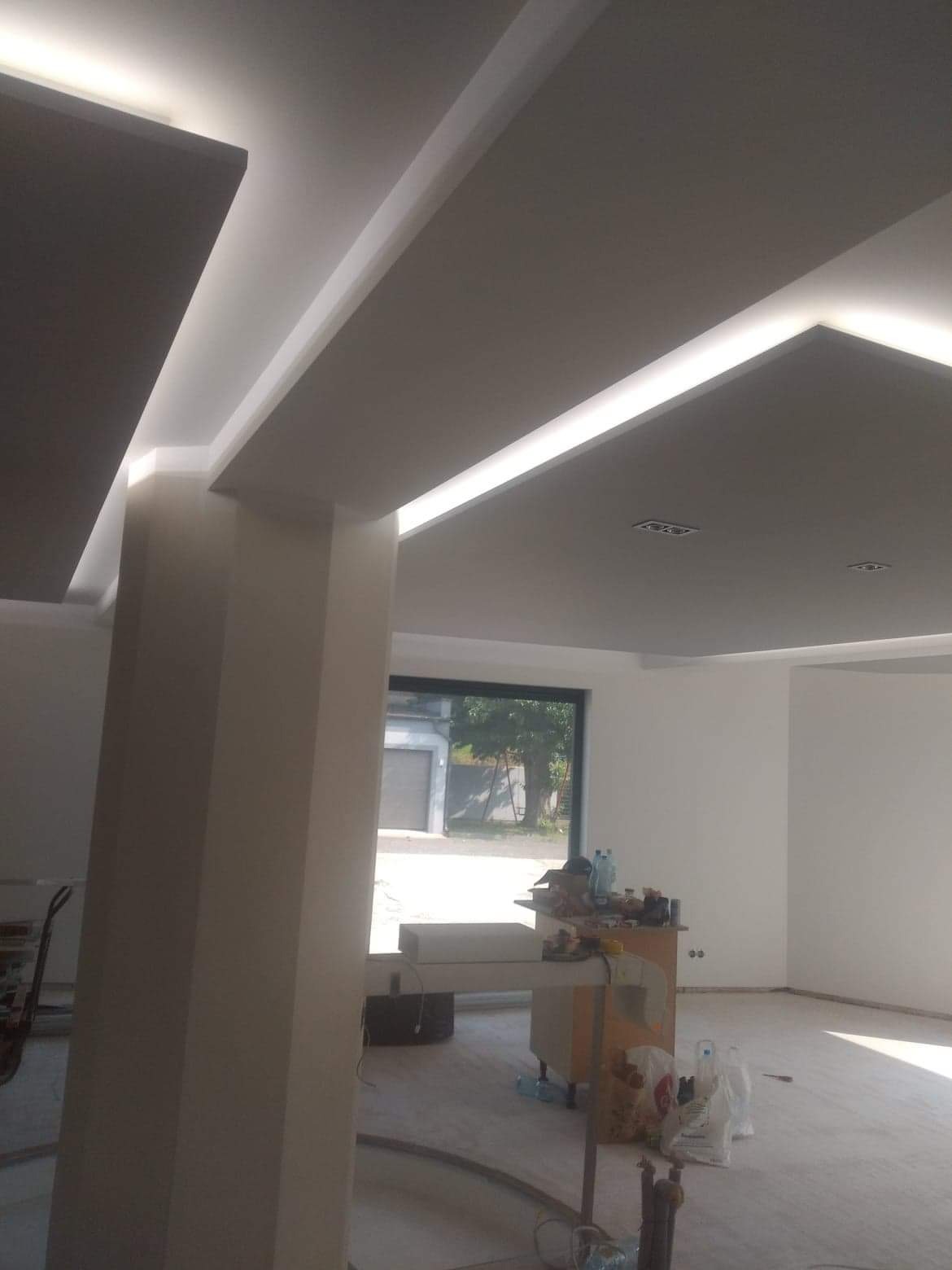 Nowoczesne oświetlenie sufitowe LED w jasnym, przestronnym wnętrzu, widok na filar i okno z widokiem na zewnątrz, podłoga częściowo zabezpieczona folią malarską.