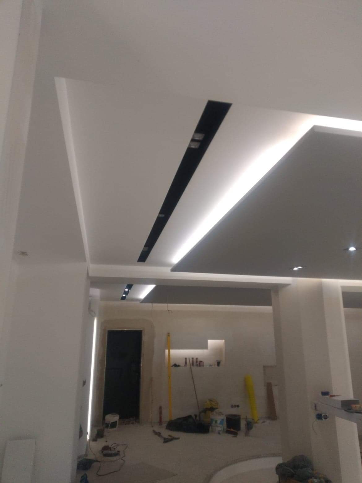 Nowoczesny sufit podwieszany z czarnymi wstawkami i oświetleniem LED, widok na pomieszczenie w trakcie prac wykończeniowych, z widocznymi narzędziami i materiałami budowlanymi.