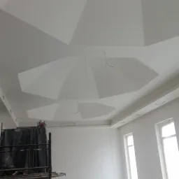 Białe wnętrze w trakcie remontu z dekoracyjnym sufitem z płyt karton gips, widoczne rusztowanie, kable wystające z sufitu i ślady po pracy na podłodze.