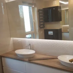 Nowoczesna łazienka z dwiema owalnymi umywalkami nablatowymi, drewnianym blatem i białymi szafkami, widoczna w lustrze kabina prysznicowa i grzejnik drabinkowy.