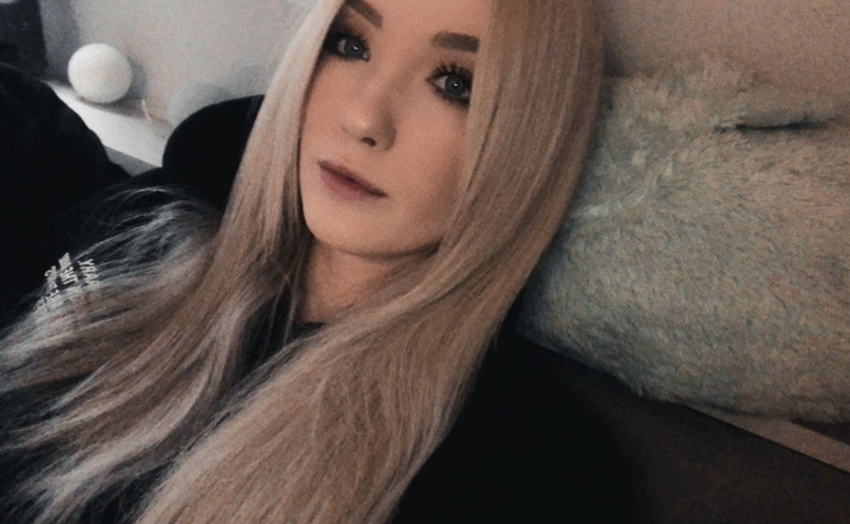 Portret młodej kobiety z długimi, blond włosami, leżącej na miękkiej, futrzanej poduszce, z widoczną kulą świetlną w tle.