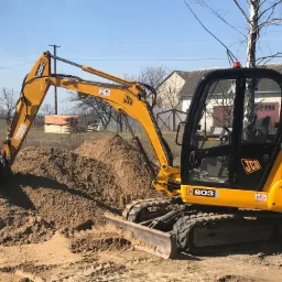 Żółta koparka JCB 803 nabiera ziemię, w tle budynek gospodarczy i ułożone deski.