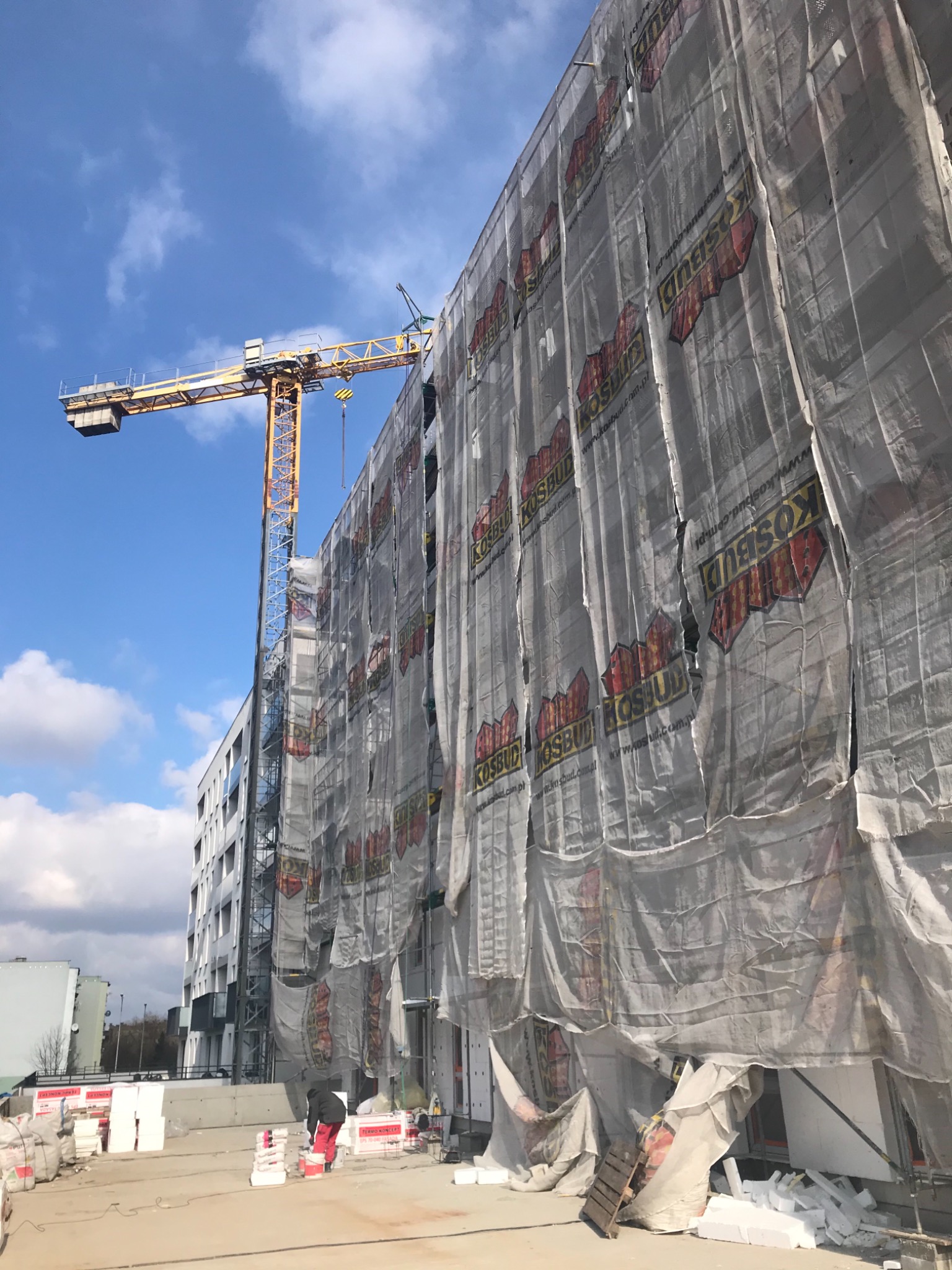 Budynek w trakcie budowy osłonięty siatką z logo firmy budowlanej, widoczny żuraw budowlany na tle błękitnego nieba z chmurami, na placu budowy materiały izolacyjne i pracownik w czerwonych spodniach.