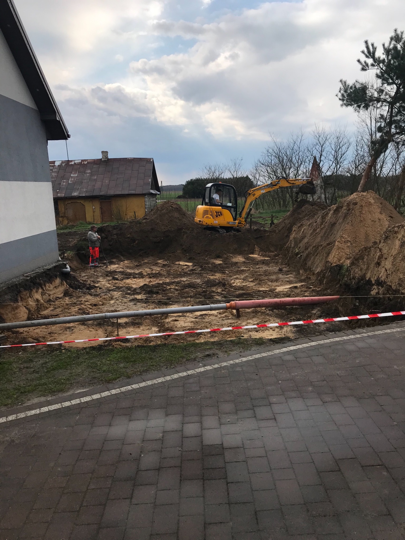 Żółta koparka JCB wykonuje wykop w ziemi, widoczne rury w wykopie, pracownik w pomarańczowych spodniach nadzoruje prace, teren zabezpieczony taśmą ostrzegawczą, w tle budynki.