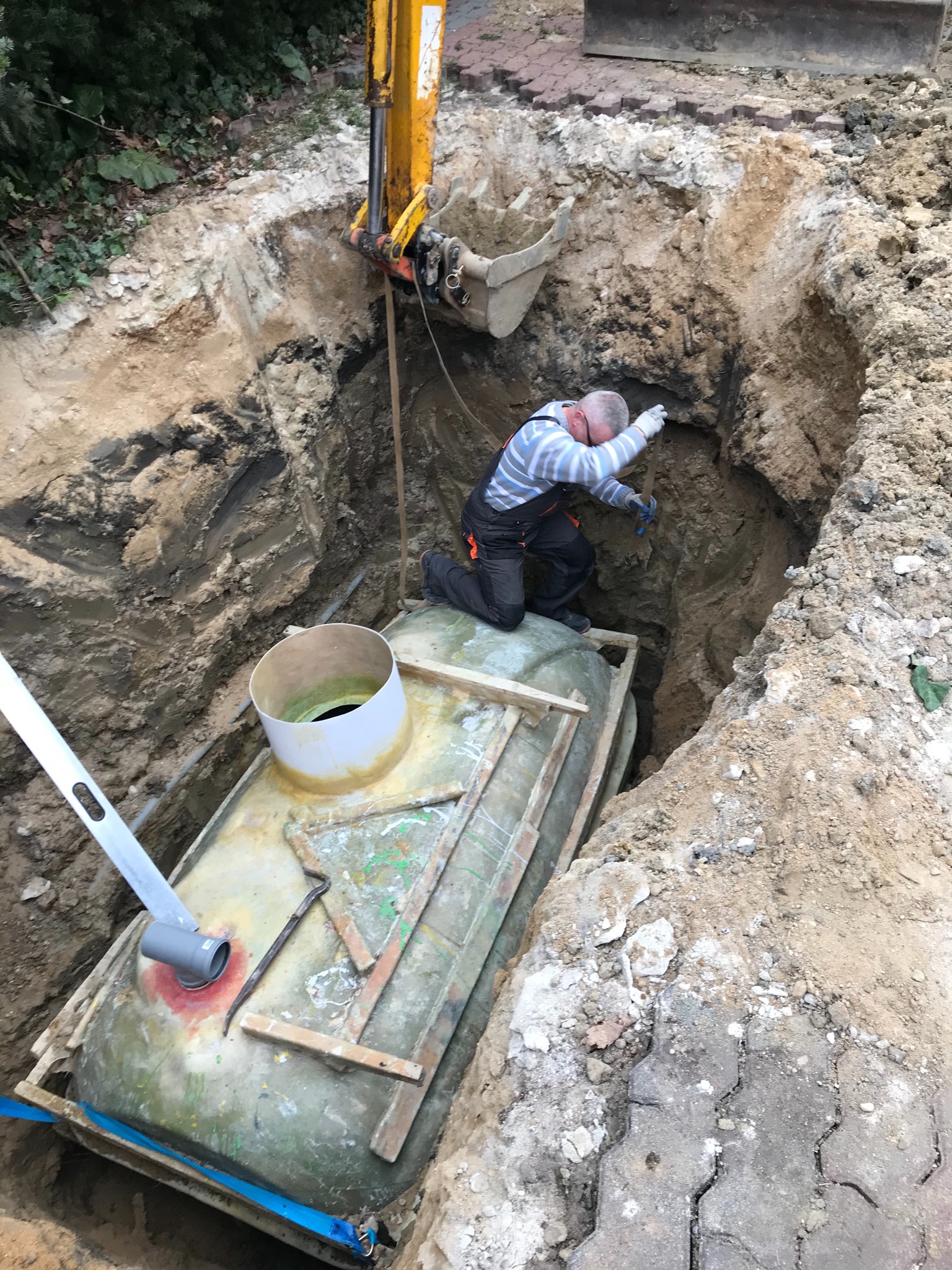 Instalacja szamba: pracownik w kombinezonie umieszcza zbiornik w wykopie za pomocą koparki, widoczny poziomica i elementy przyłączeniowe.