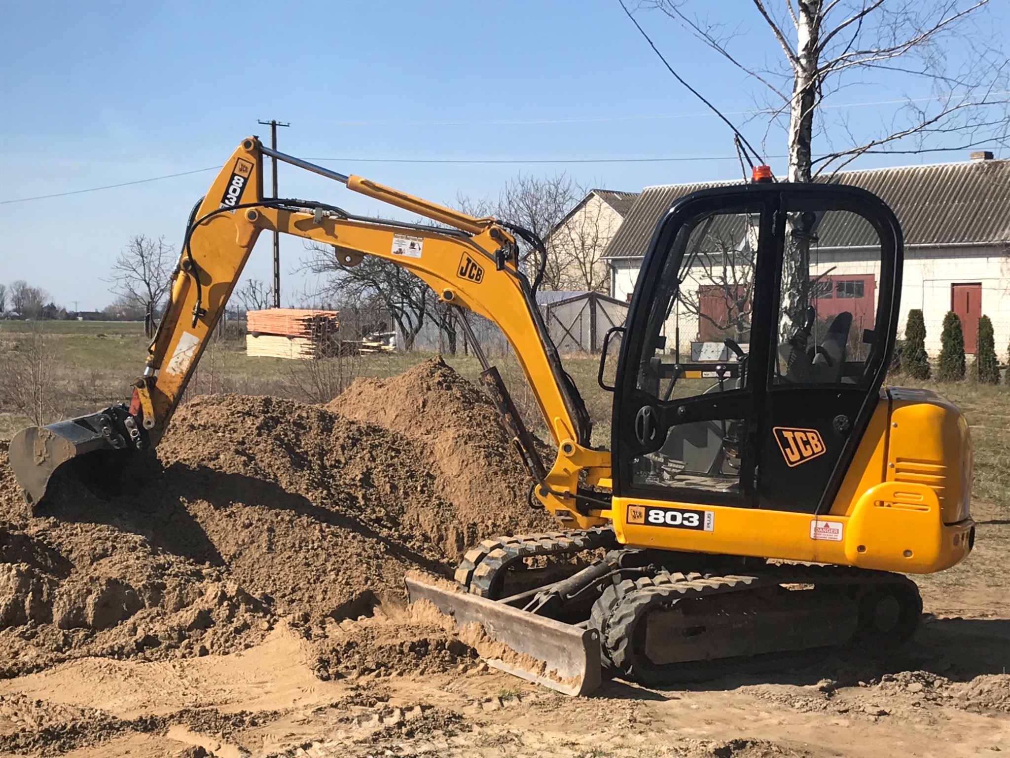 Żółta koparka JCB 803 nabiera ziemię, w tle budynek gospodarczy i ułożone deski.