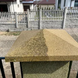 Betonowy słupek ogrodzeniowy z piramidalnym daszkiem, widoczna różnica w czystości połowy powierzchni, zarysowania i zabrudzenia, w tle fragment ogrodzenia posesji.