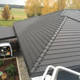 Ciemnoszary dach z dachówką karpiówką, widok z góry, na tle jesiennego krajobrazu i samochodu serwisowego z otwartym bagażnikiem, w którym widoczny jest sprzęt do pracy.