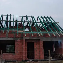 Budowa domu jednorodzinnego w trakcie wznoszenia więźby dachowej z zielonych elementów, widoczne ślady po zaprawie na cegłach i pustakach