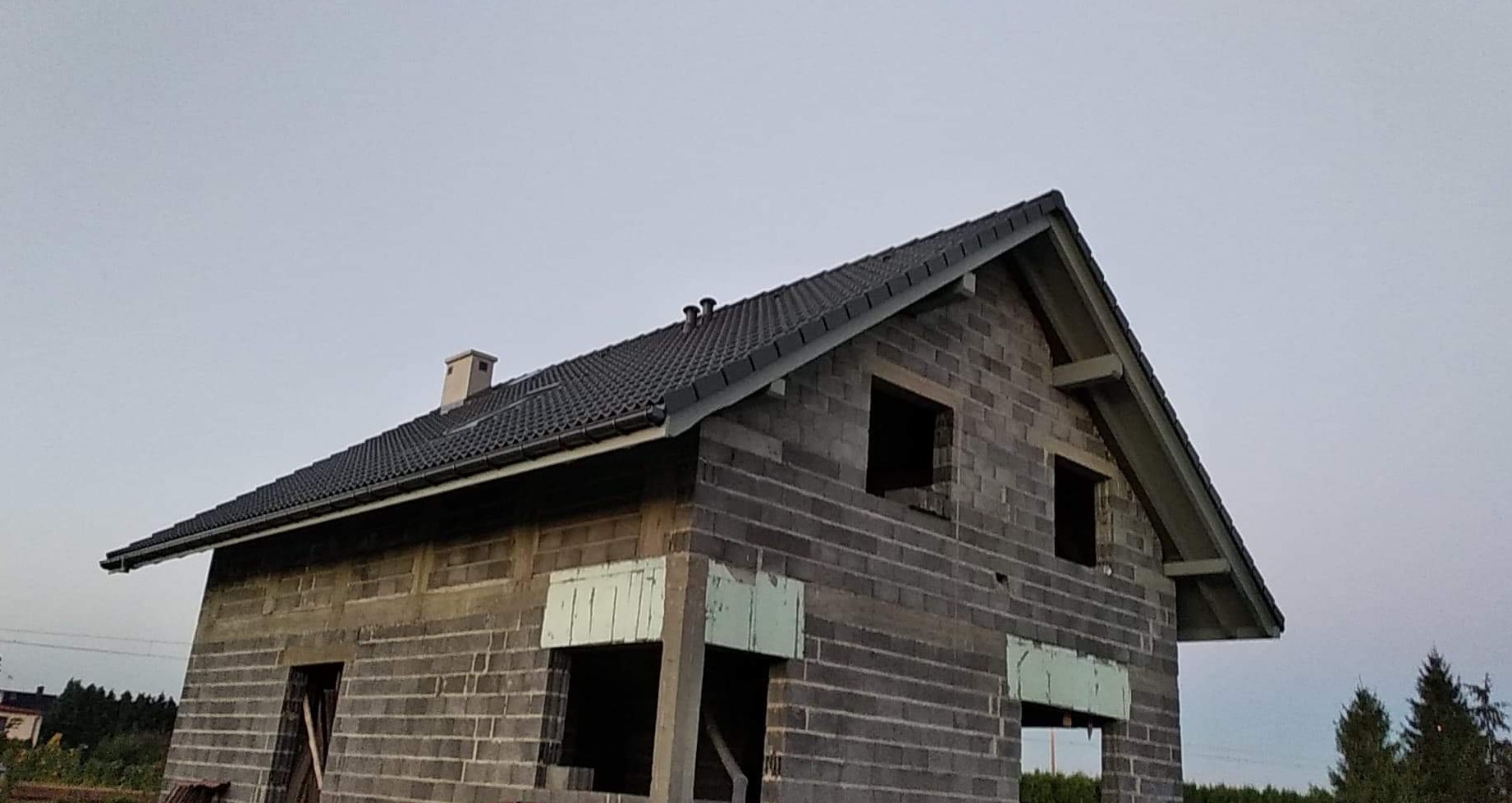 Surowy budynek w trakcie budowy z ciemną dachówką, widoczne otwory okienne i komin.