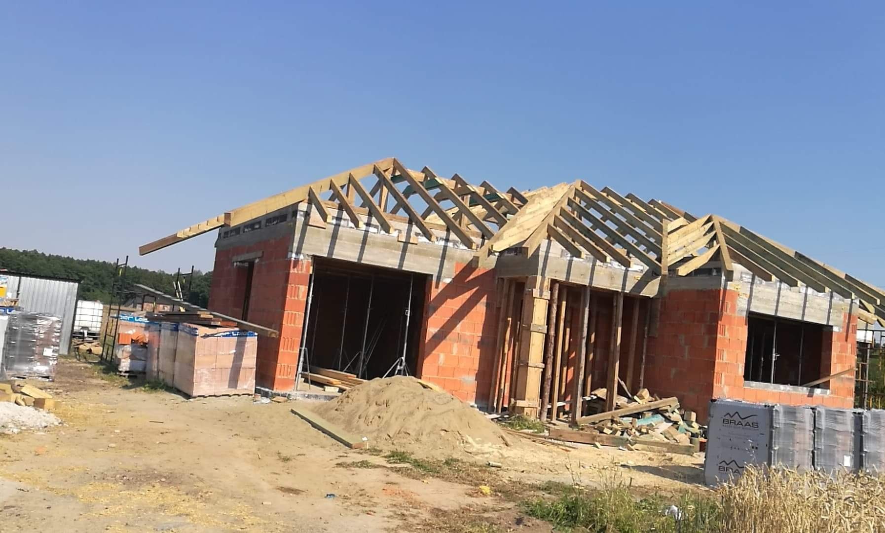 Budowa domu jednorodzinnego w stanie surowym otwartym, widoczna murowana konstrukcja z czerwonej cegły i drewniana konstrukcja dachu w trakcie montażu, z materiałami budowlanymi składowanymi obok.