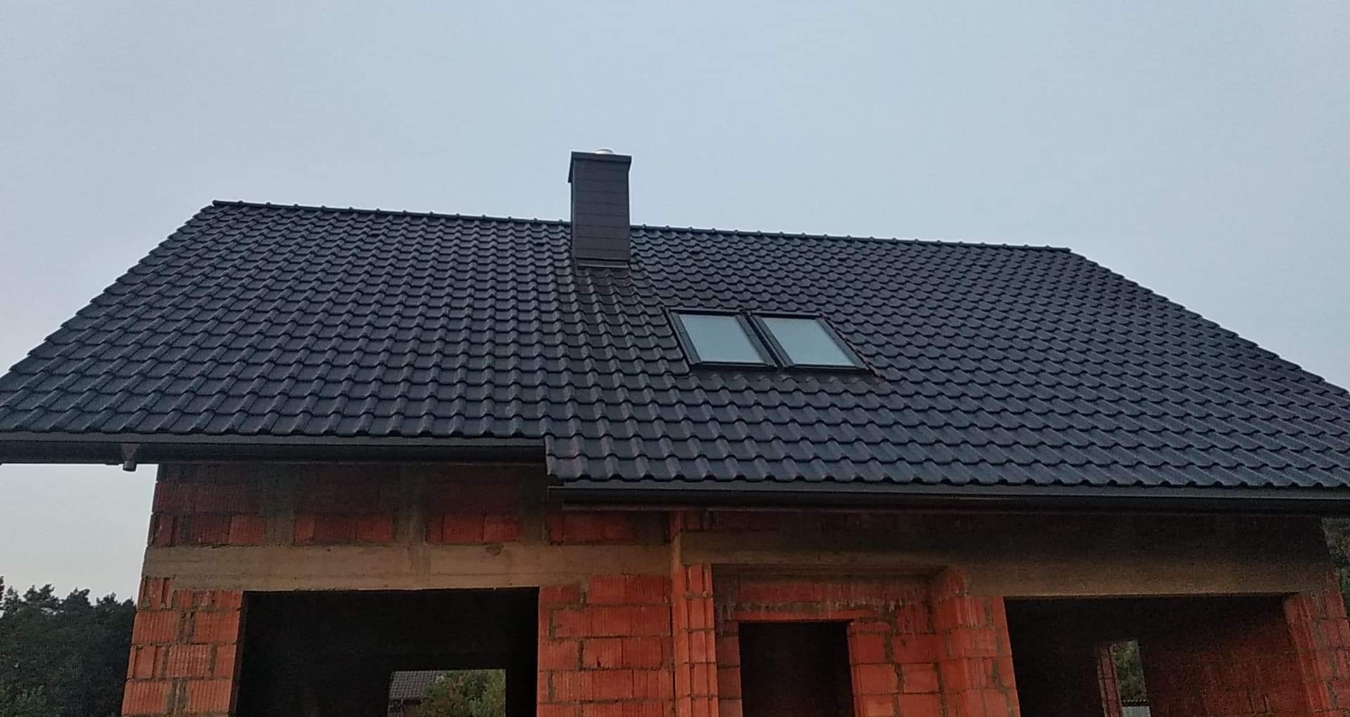 Czarny dach z dachówki modułowej z dwoma oknami dachowymi i kominem na surowym budynku z czerwonej cegły w Bojszowach.
