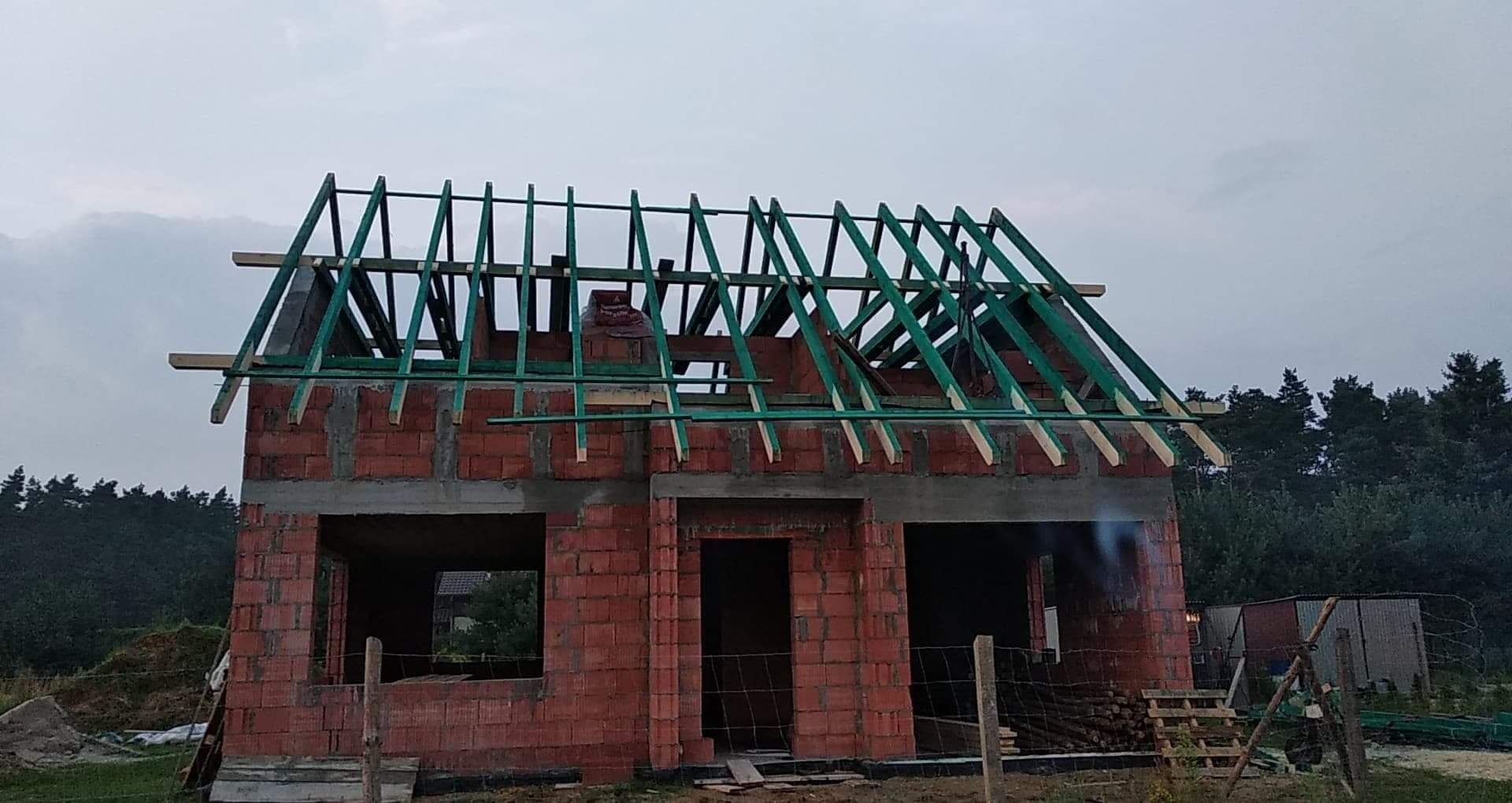 Budowa domu jednorodzinnego w trakcie wznoszenia więźby dachowej z zielonych elementów, widoczne ślady po zaprawie na cegłach i pustakach