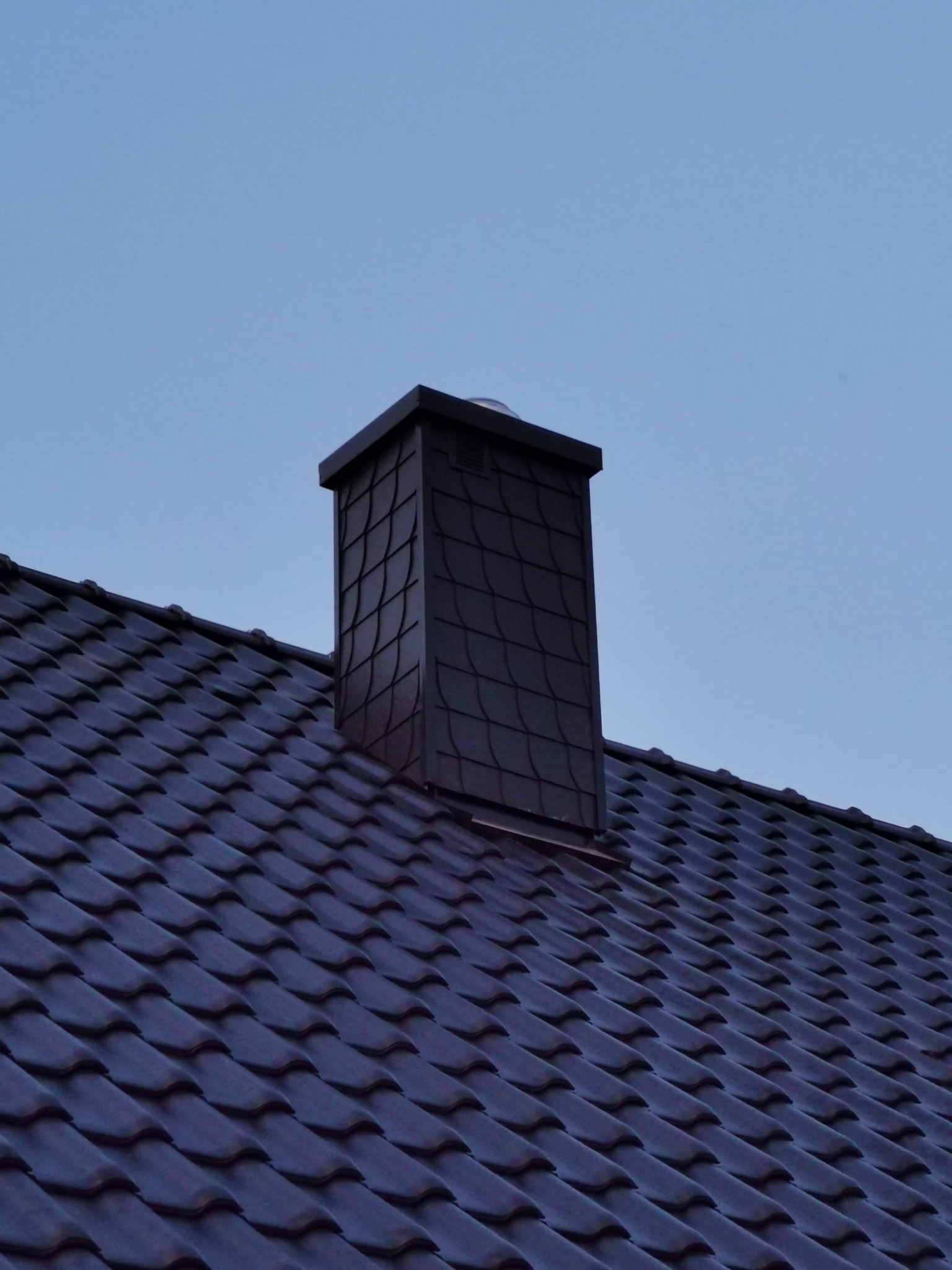 Ciemna dachówka ceramiczna z dopasowanym kolorystycznie kominem z elementami dekoracyjnymi w kształcie łusek, na tle błękitnego nieba.