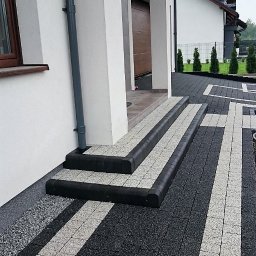 Marbruk - Ułożona kostka brukowa w dwóch kolorach prowadząca do schodów wejściowych domu z ciemnymi obrzeżami, widoczne elementy elewacji budynku i trawnik z tujami w tle.