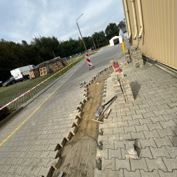 Marbruk - Wykopany rów w kostce brukowej, przygotowany do ułożenia nowej warstwy, widoczne narzędzia: łopata i kielnia, palety w tle.