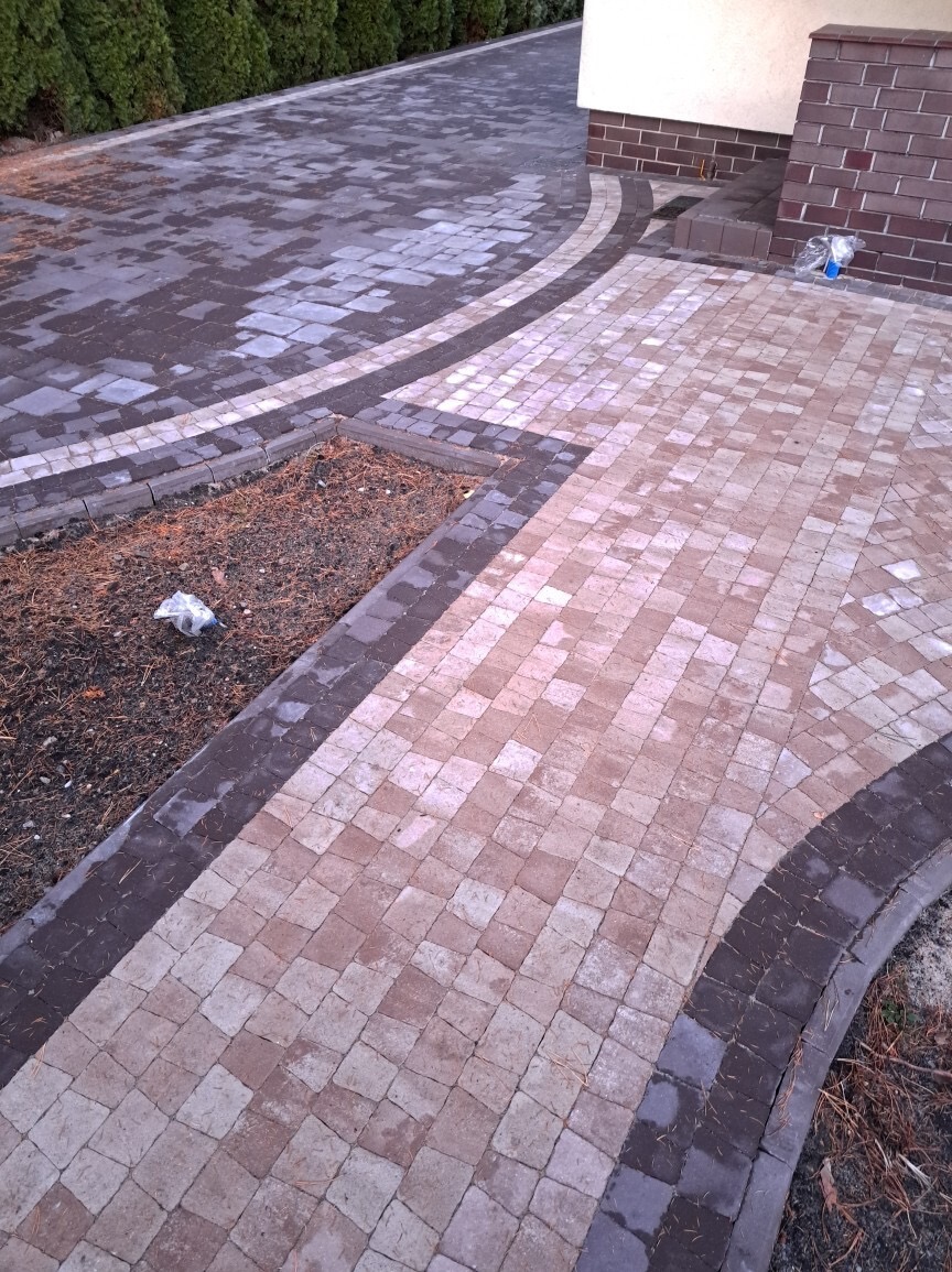 Ułożona nawierzchnia z kostki brukowej w dwóch odcieniach szarości i beżu, z obrzeżem w ciemniejszym kolorze, tworząca zakrzywiony wzór przy budynku z cegły.