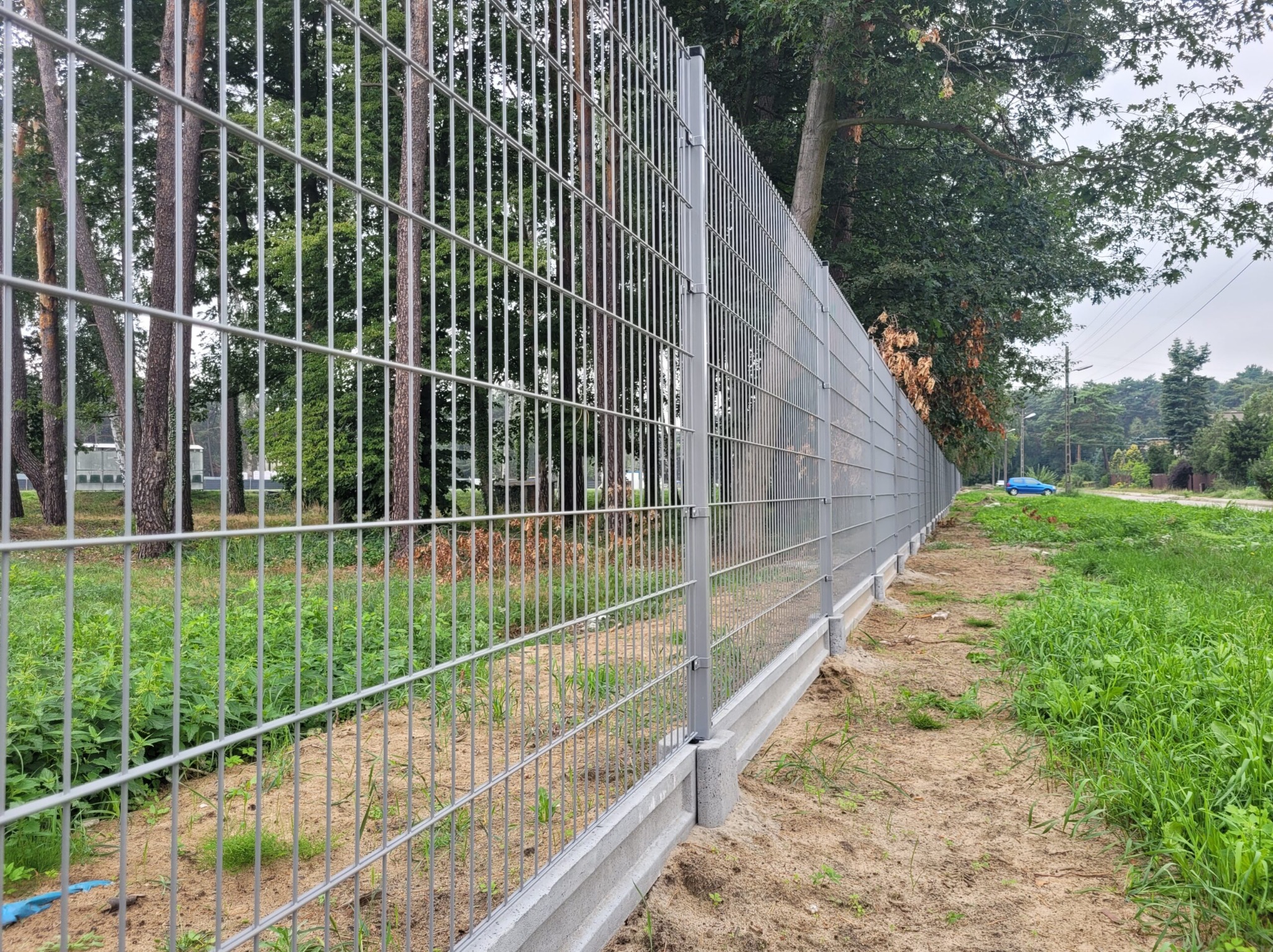 Długa, prosta linia szarego, metalowego ogrodzenia panelowego z podmurówką betonową, rozciągająca się wzdłuż drogi z zielenią w tle.