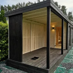 Nowoczesna, drewniana konstrukcja z ciemnym obramowaniem, prawdopodobnie w trakcie budowy, otoczona zieloną siatką i kamieniami, widoczna wewnętrzna część z jasnego drewna i przeszklone drzwi.