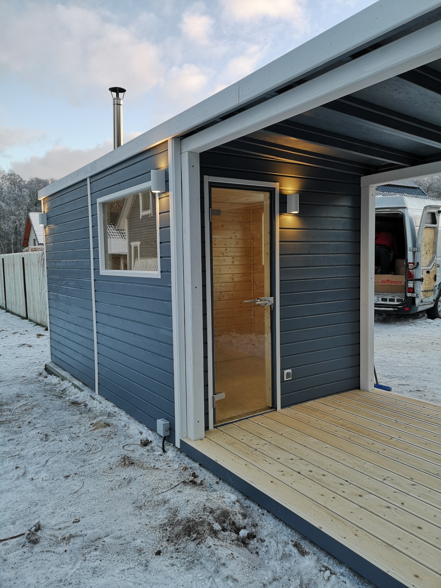 Zewnętrzna, drewniana kabina z przeszklonymi drzwiami i kominem na dachu, oświetlona kinkietami, z drewnianym tarasem, otoczona śniegiem, z częściowo widocznym samochodem dostawczym w tle.