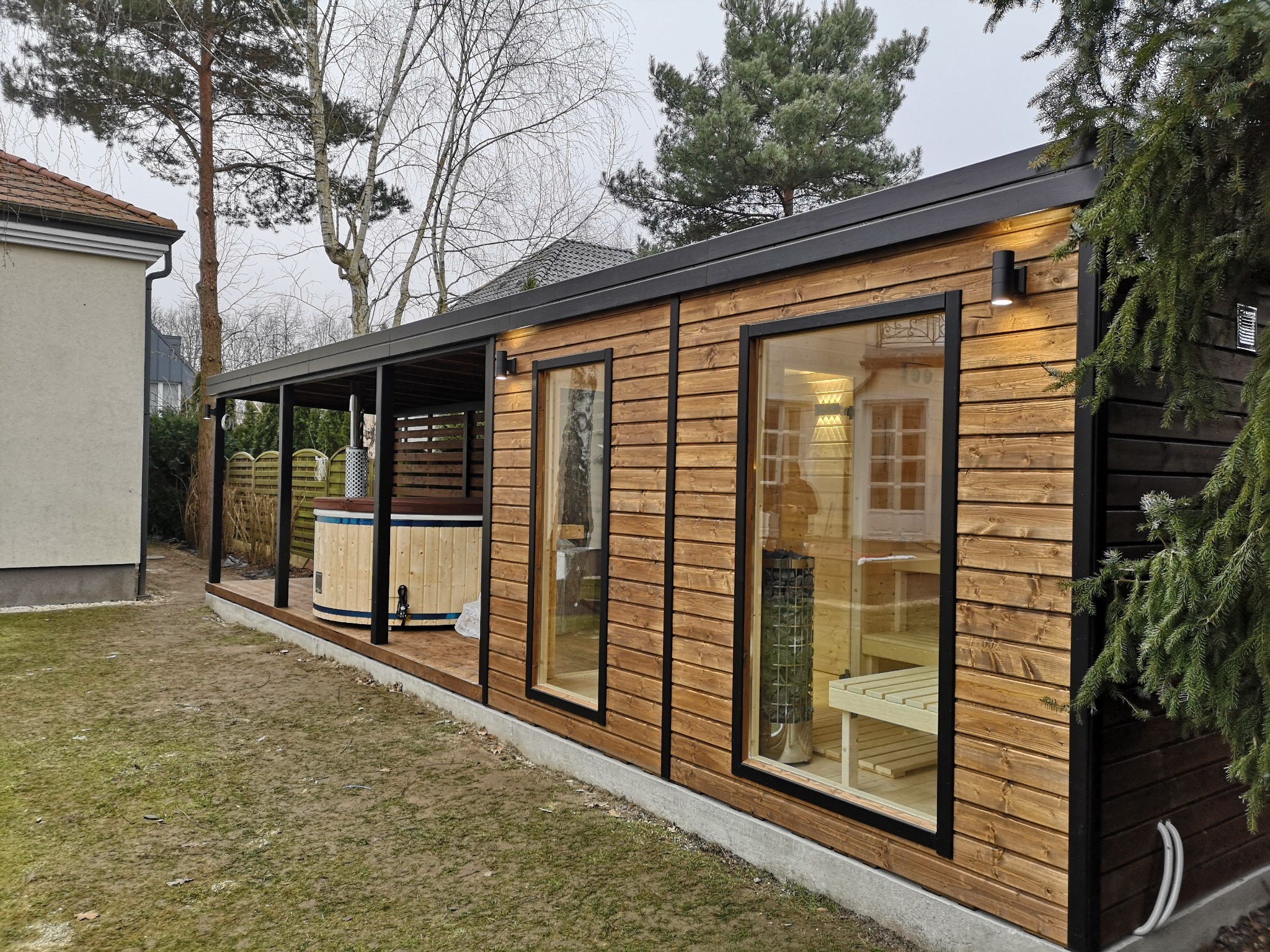 Drewniana konstrukcja z sauną i balią na zewnątrz, widoczna przez okna wewnętrzna aranżacja sauny z ławkami i piecem, obok balia z metalowym piecem pod zadaszeniem.
