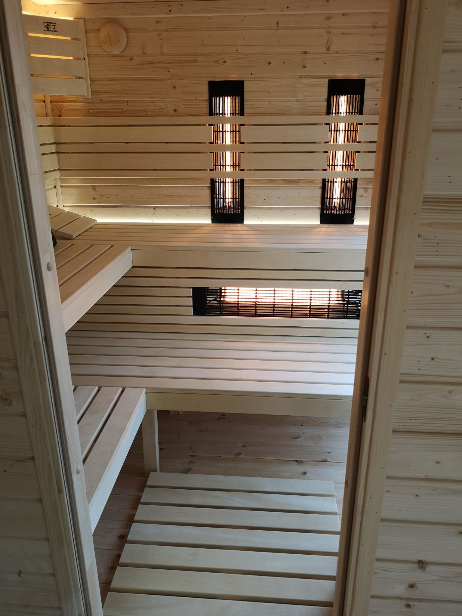Wnętrze drewnianej kabiny z podświetlanymi panelami grzewczymi, widok z zewnątrz przez uchylone drzwi.