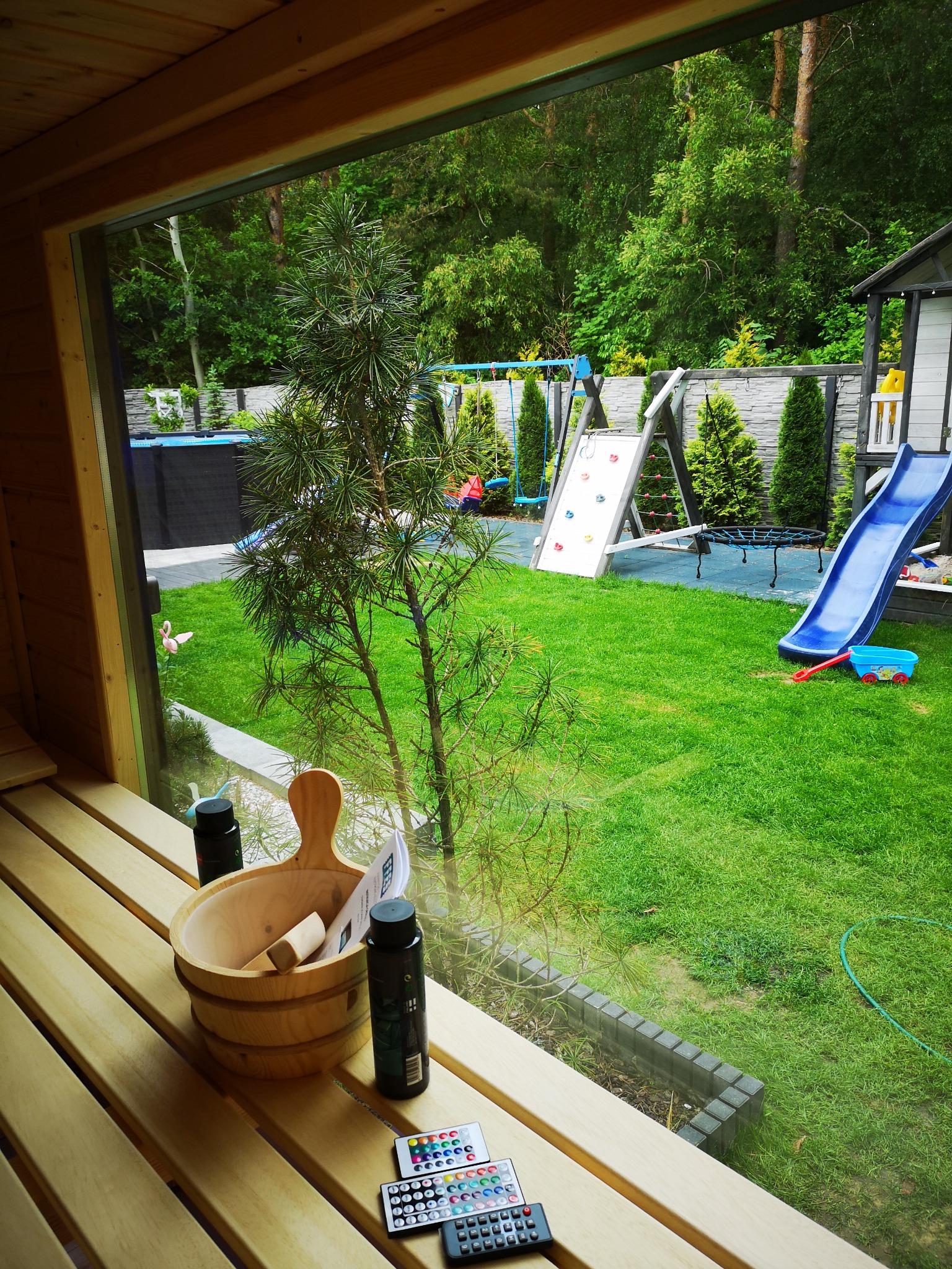 Wnętrze drewnianej kabiny z widokiem na zielony ogród z placem zabaw, wiadro i akcesoria do pielęgnacji na ławce.
